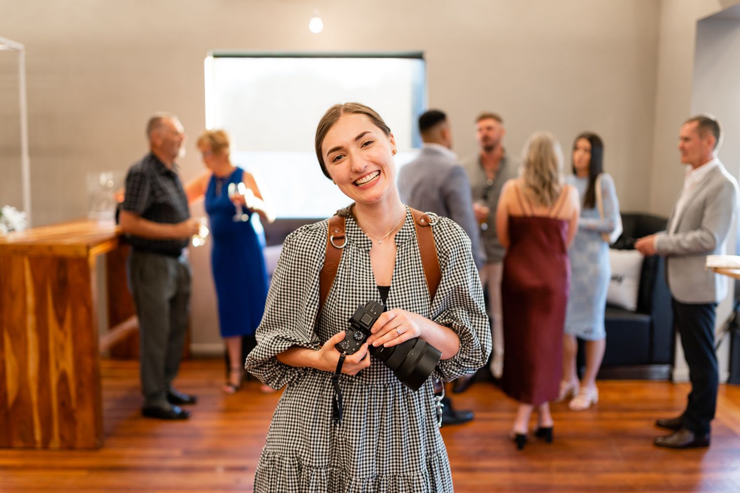 Event photographer in a grey dress standing in a room with people mingling in the background.