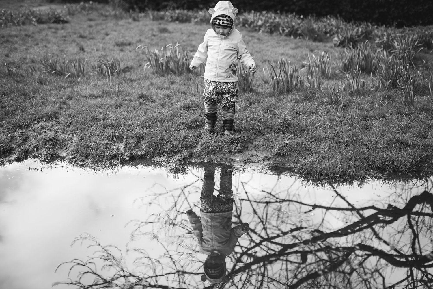 Small child reflected in puddle while standing in grassy field.