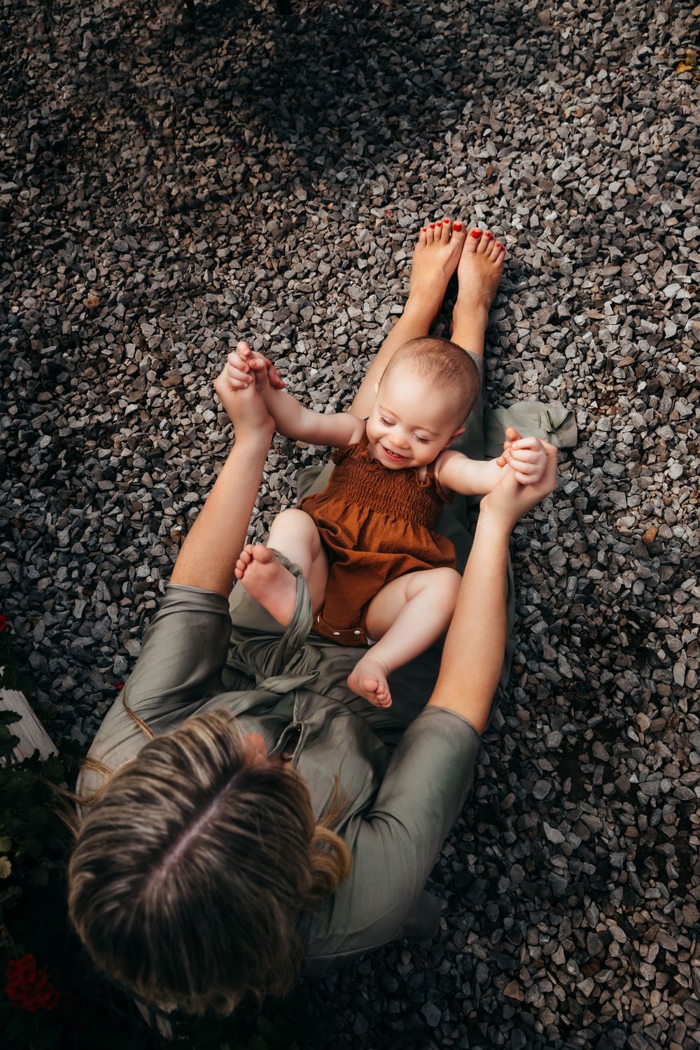 Parent lying on gravel ground playing and holding up a smiling baby wearing an orange romper.