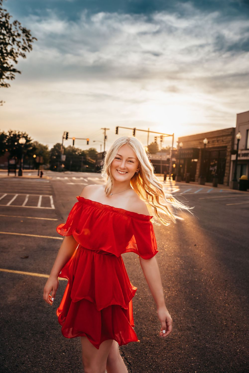 A person in a red off-shoulder dress stands on a street at sunset with golden light and moody clouds overhead.