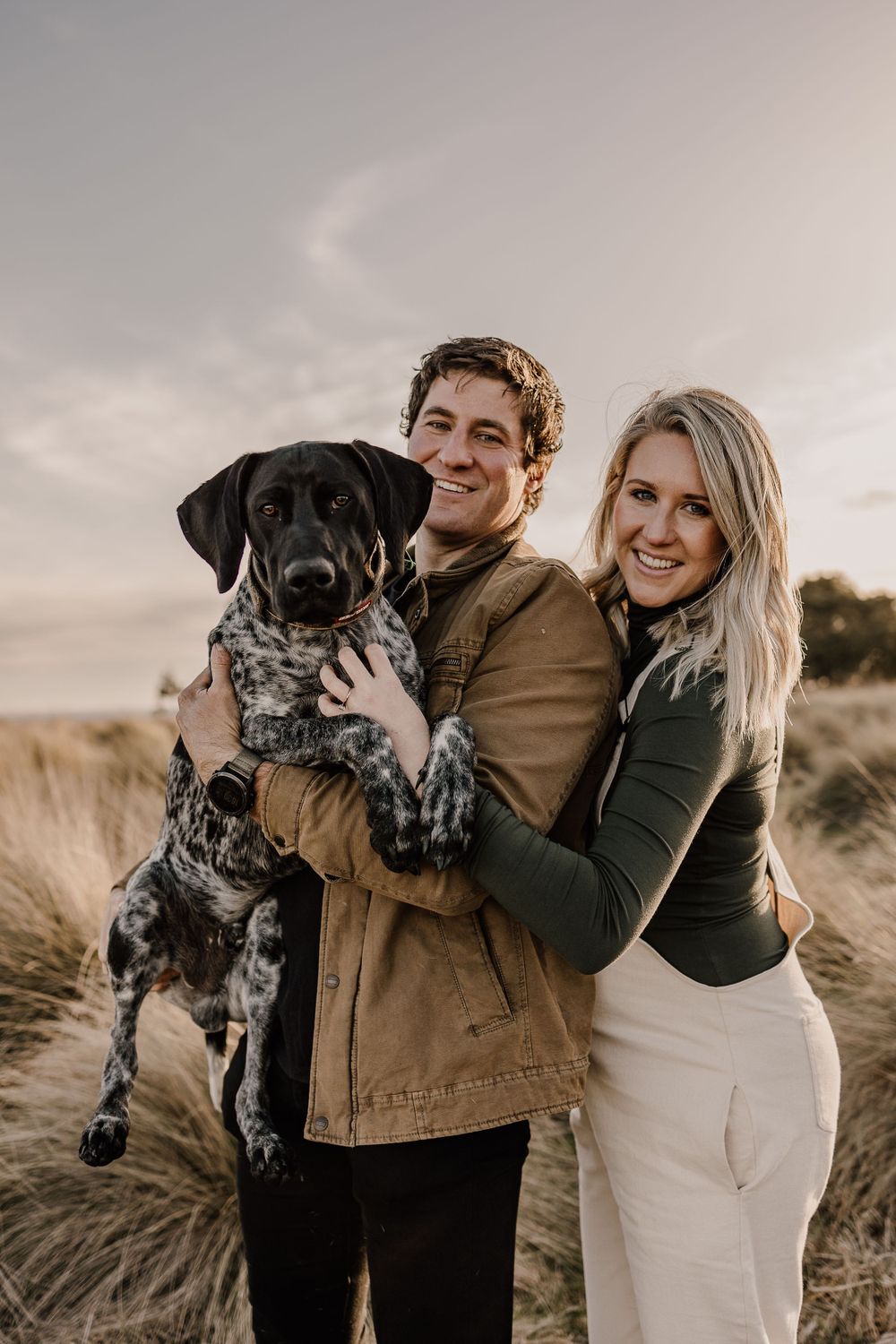 Family portrait featuring a couple with their spotted dog against a backdrop of wild grasses.