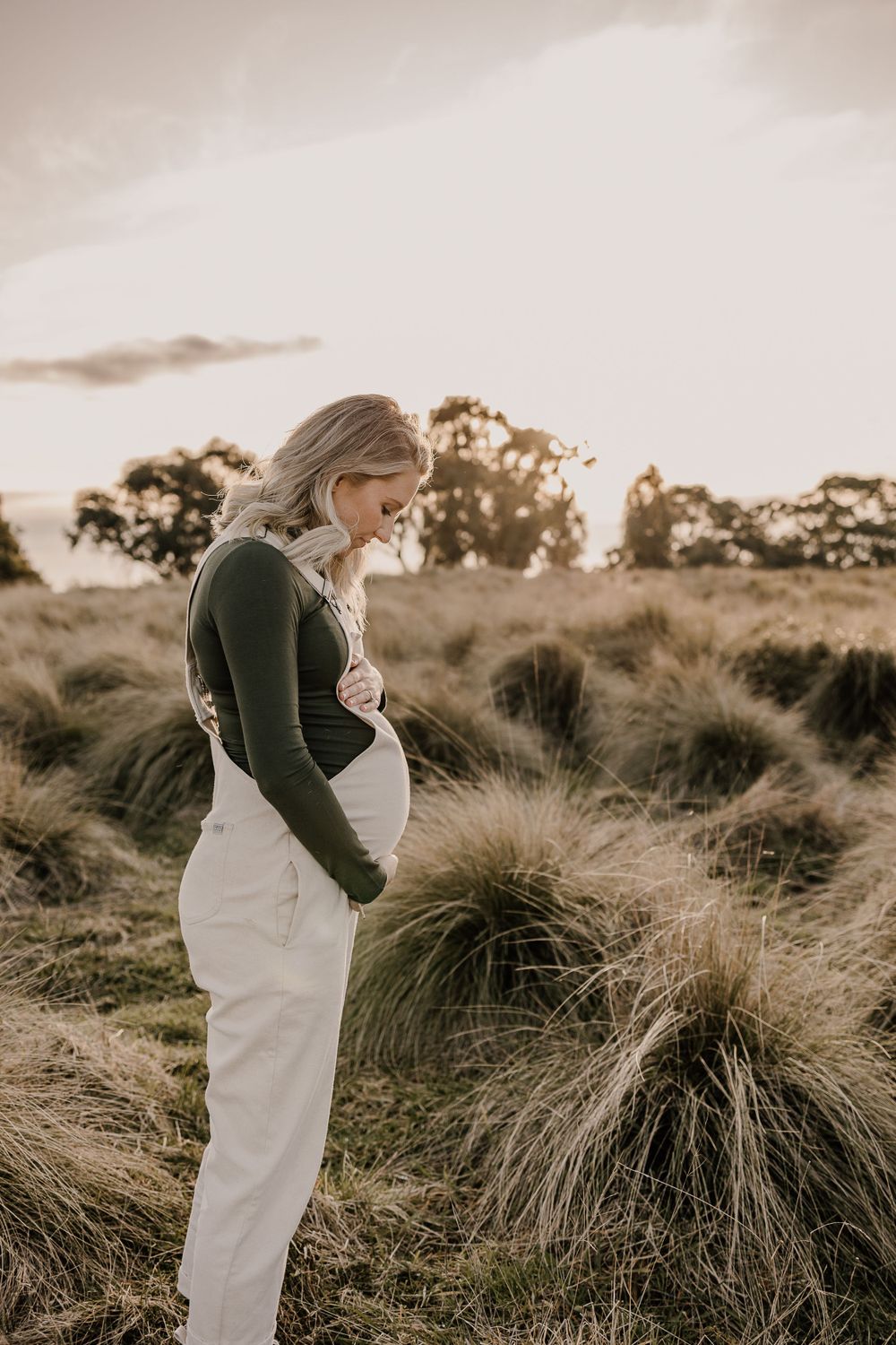A person in white overalls and dark sweater stands in tall grass at sunset.