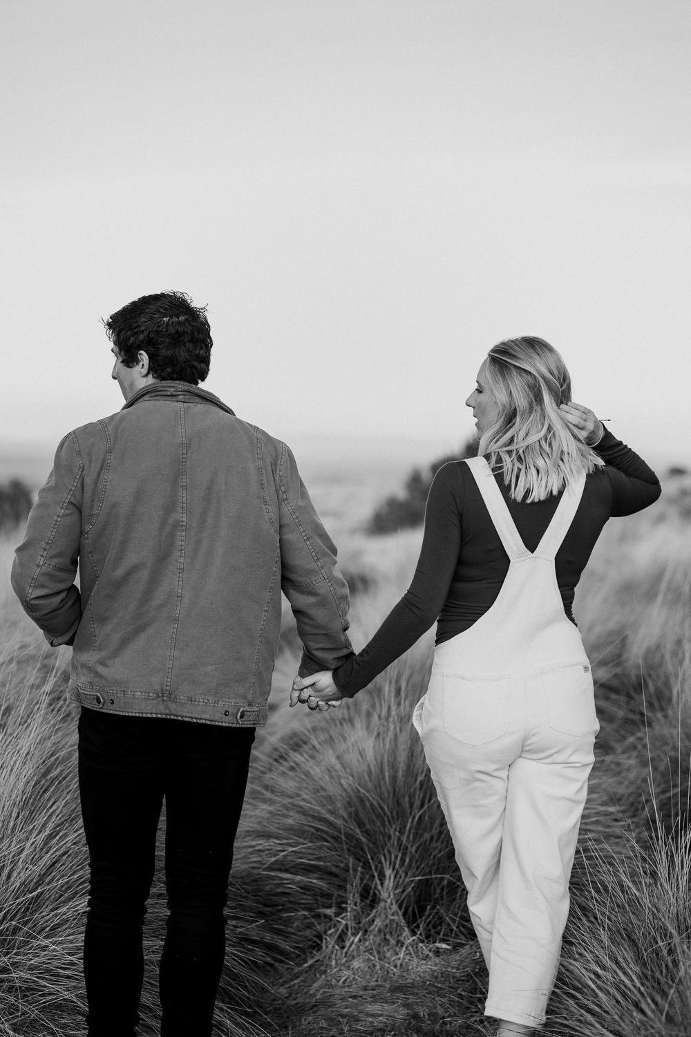 A couple holds hands while walking through tall grass in black and white.