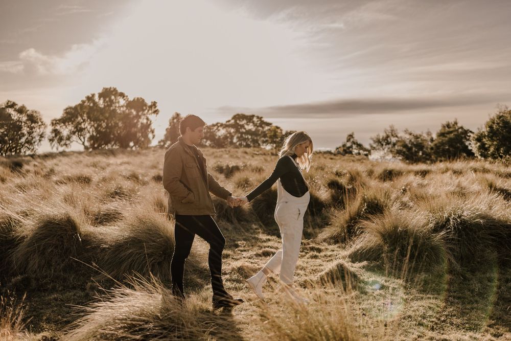 A couple walks hand in hand through tall grass during golden hour.