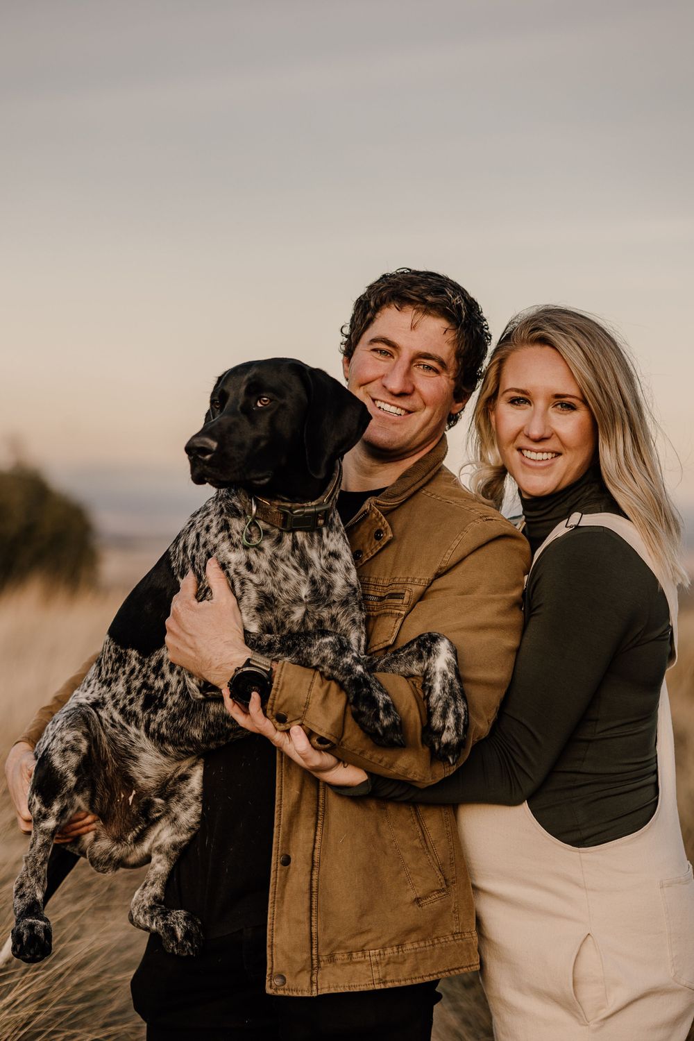 A couple poses with their spotted dog in a field during golden hour.