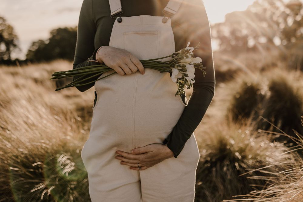 Expectant mother in white overalls holds flowers while cradling her baby bump in golden sunlight.