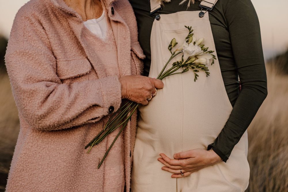 A maternity photo showing hands holding delicate wildflowers against a pregnant belly in natural light.