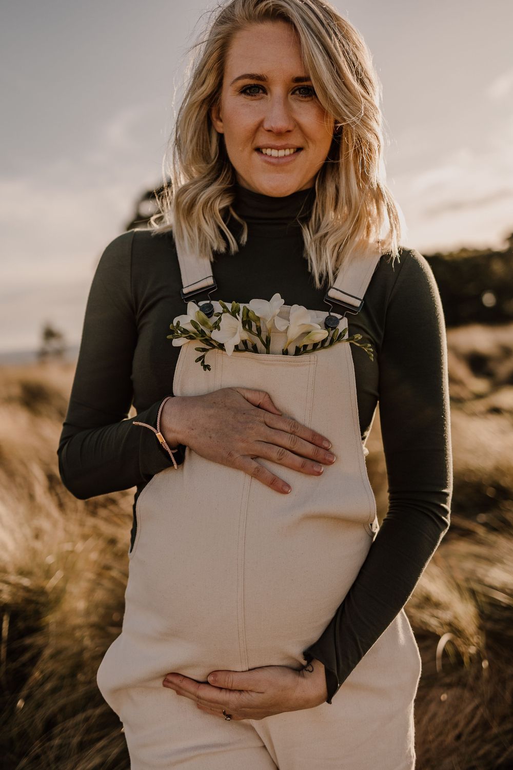 Expecting mother wearing white overalls and dark sweater holds flowers in warm evening light.