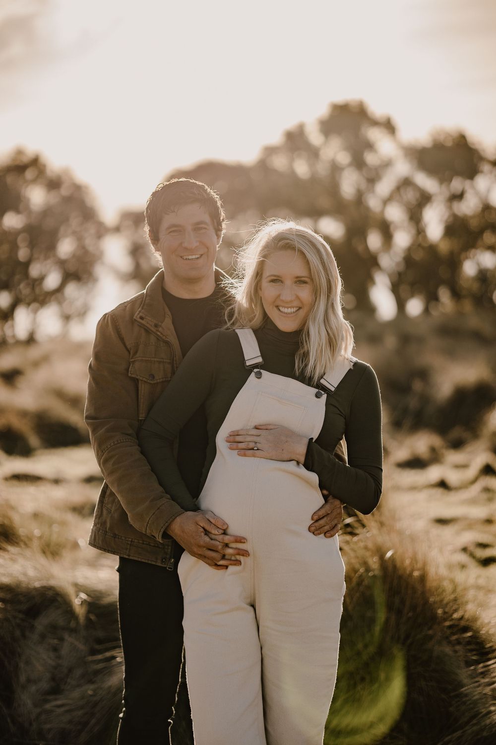 An expectant couple poses together in white overalls and brown jacket at sunset.