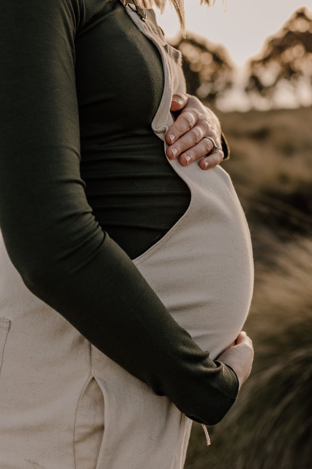 Close-up of hands cradling a pregnant belly in white overalls and dark sweater.