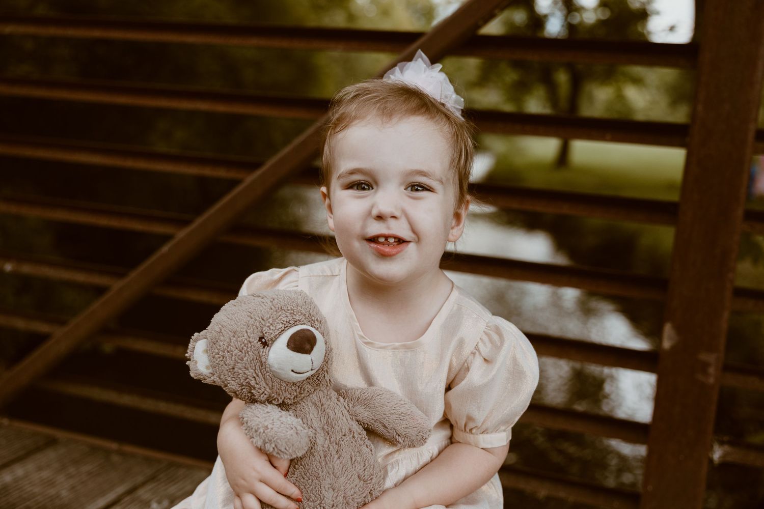 A child in a puff-sleeve dress poses with their favorite teddy bear on wooden steps.