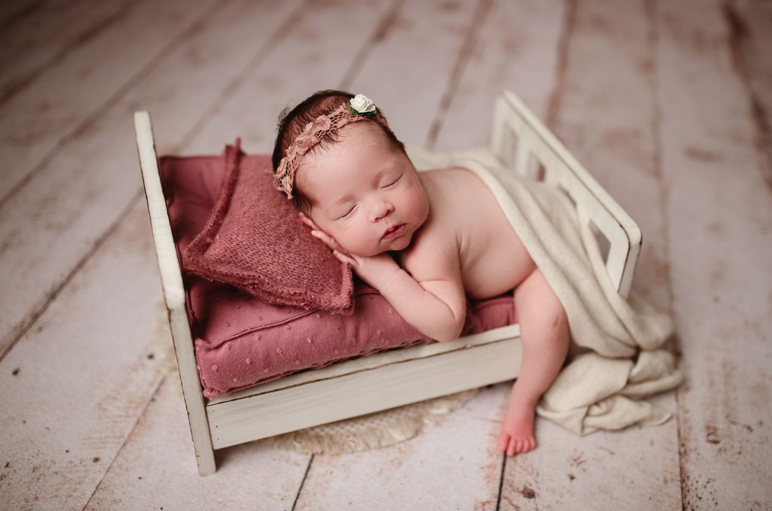 Sleeping newborn on burgundy blanket in white wooden bed.
