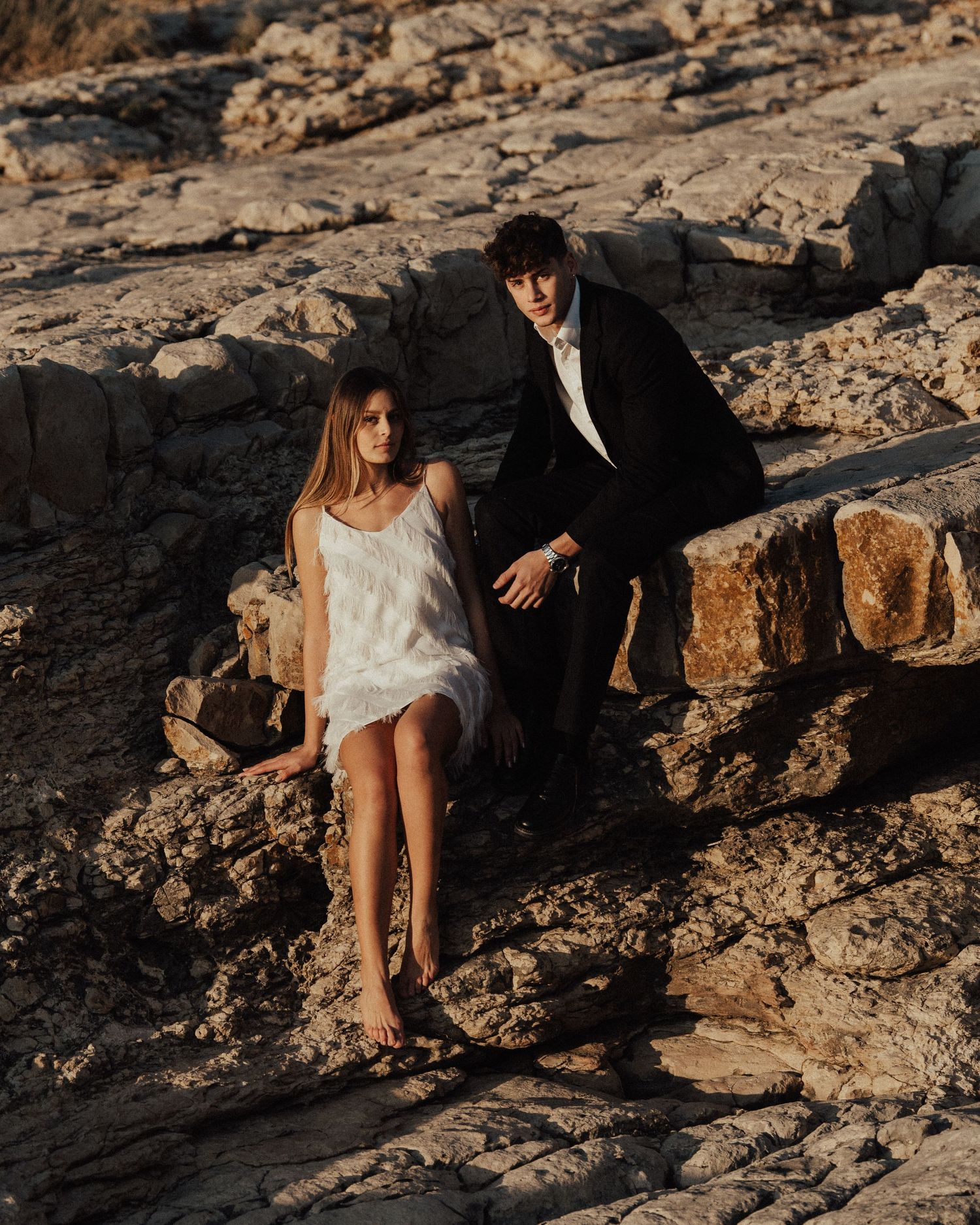 Couple sitting together on rugged coastal rocks during golden hour.