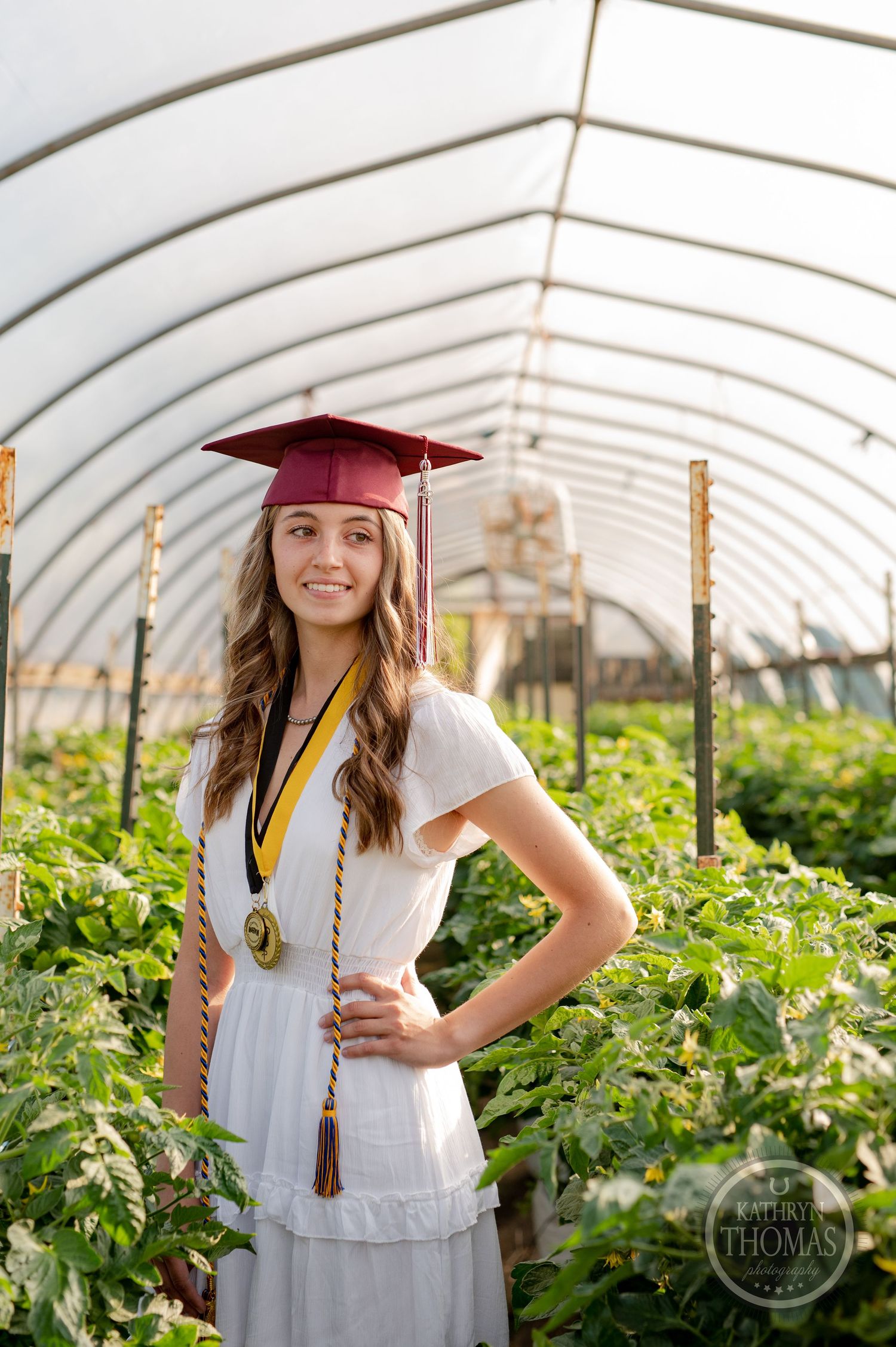 Graduate poses in white dress and maroon cap inside greenhouse filled with growing plants.