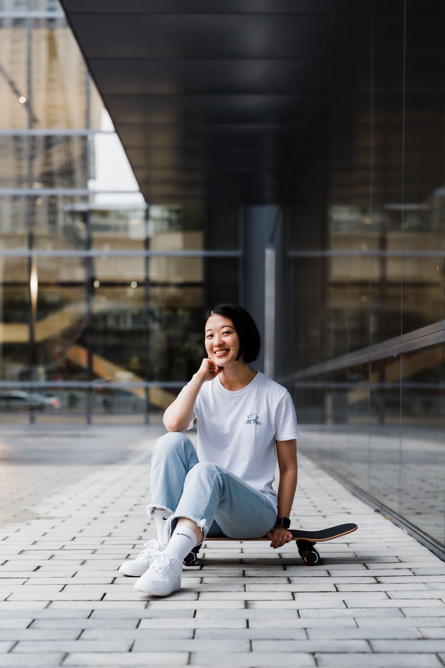 Person in casual attire sitting on a skateboard outside a modern building.