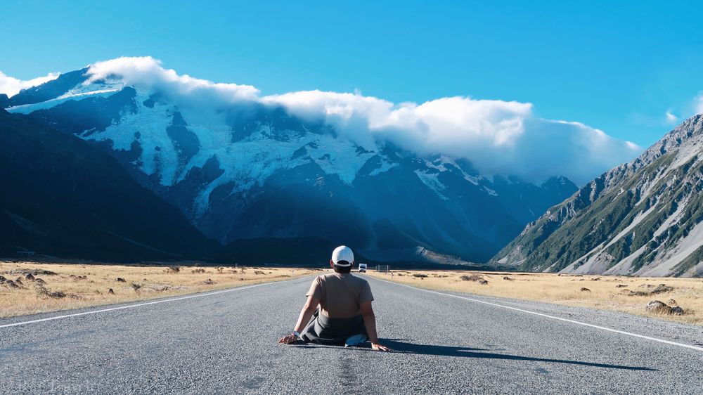 Scenic mountain road leads to snow-capped peaks under cloudy skies.