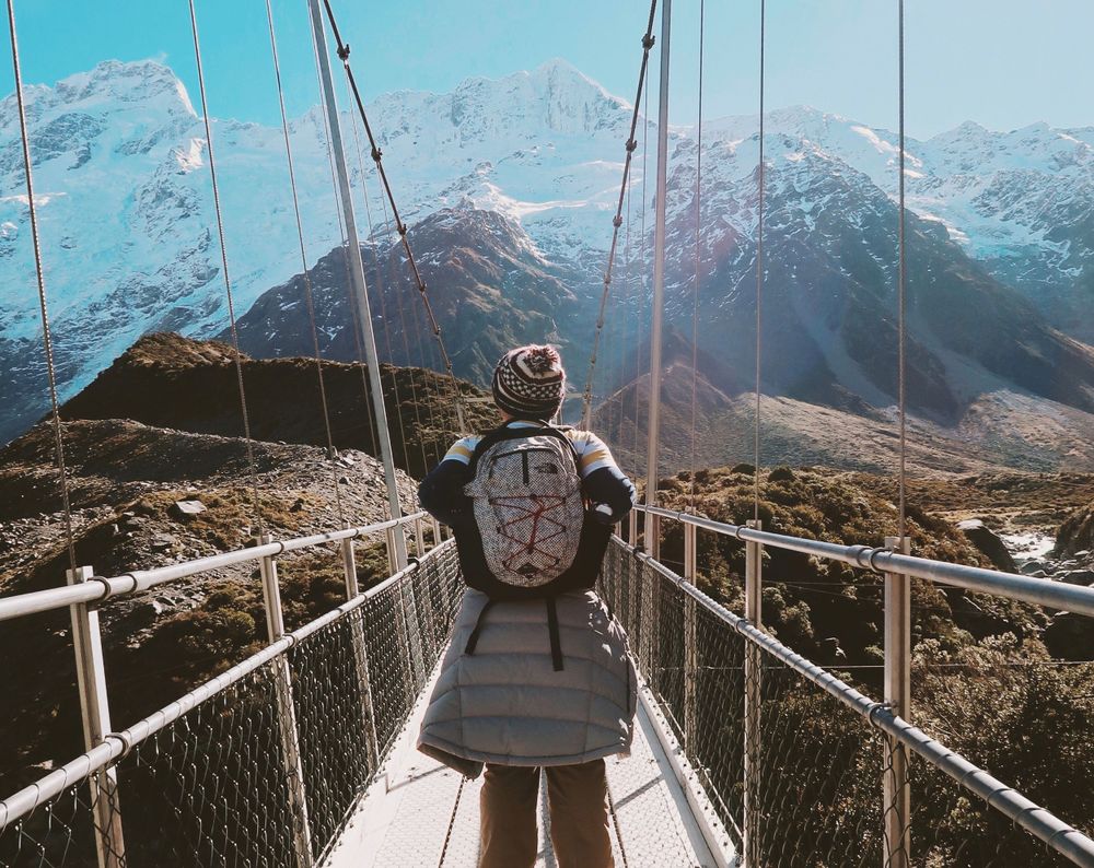 Adventure seeker crosses a suspension bridge with snow-capped mountains in the background.