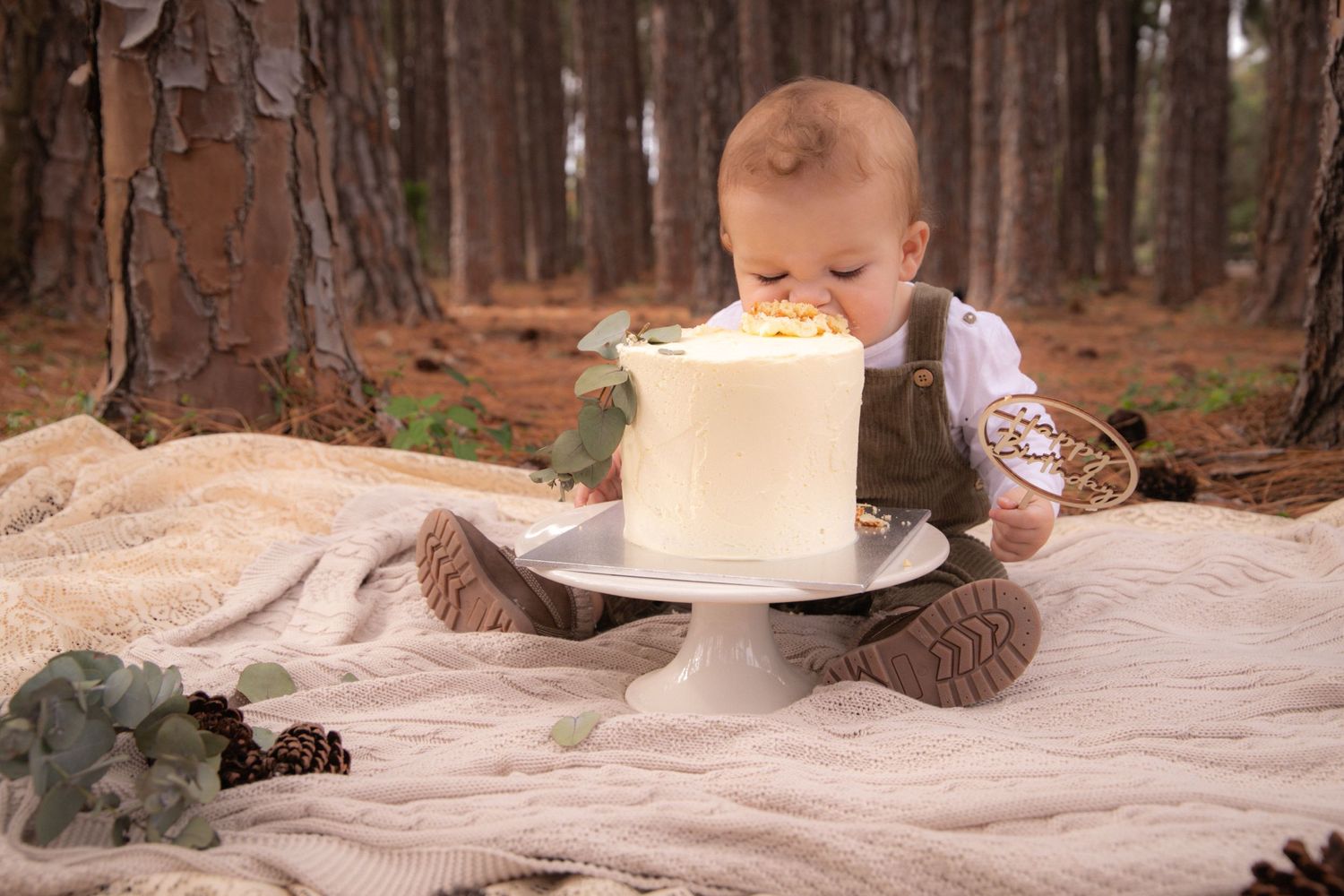 Baby leaning towards white cake on plate in forest setting with pine trees.