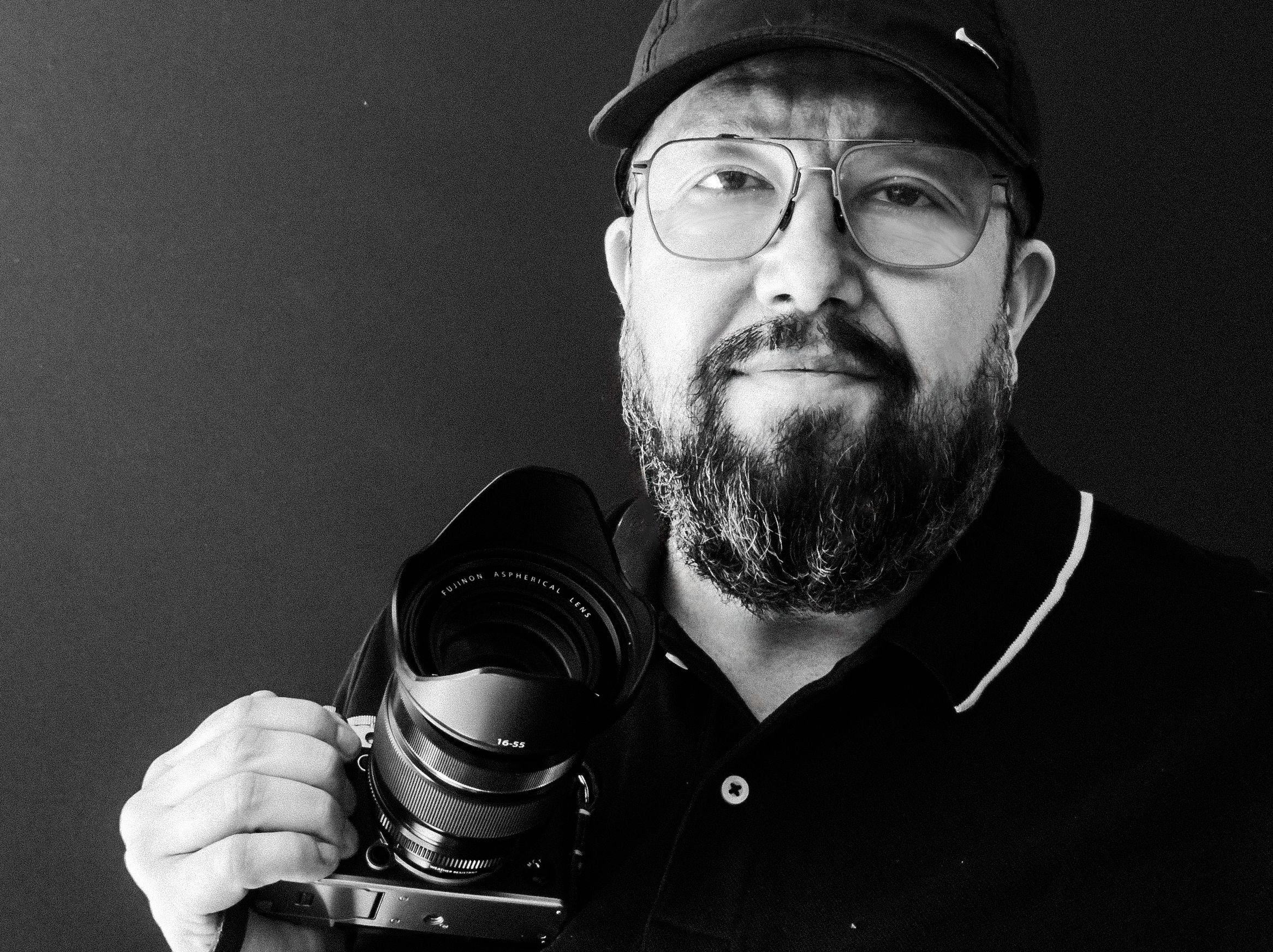 Professional photographer in black cap and glasses holding camera against dark background.