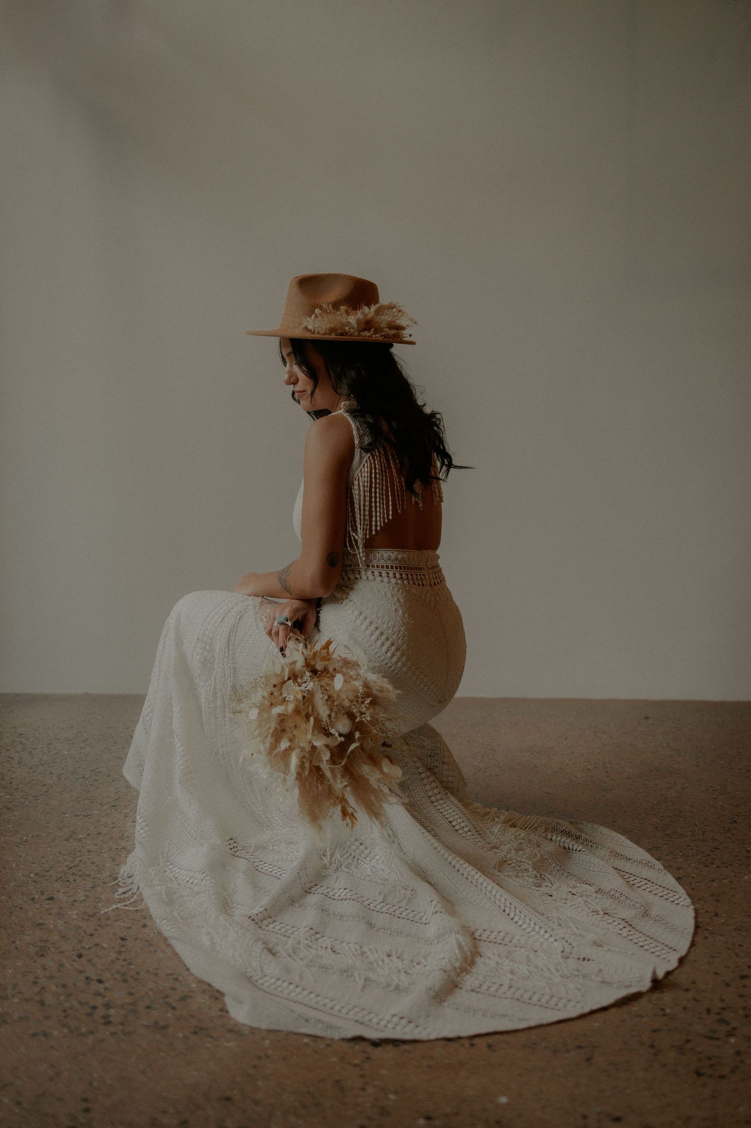 Elegant bridal portrait featuring a fitted lace gown with train and brown hat in moody lighting.