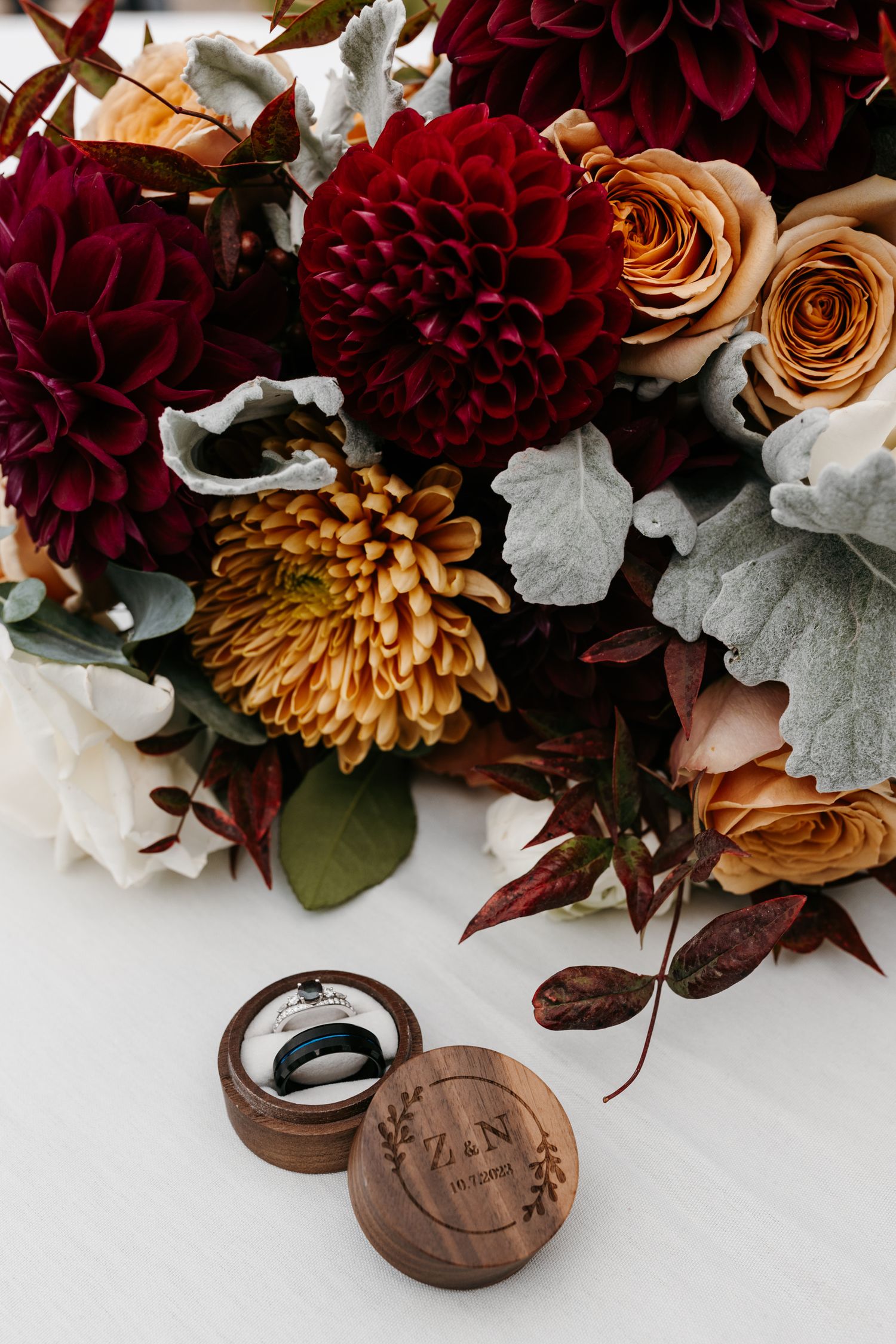 Deep red dahlias and bronze roses arranged with greenery next to wooden wedding ring box.