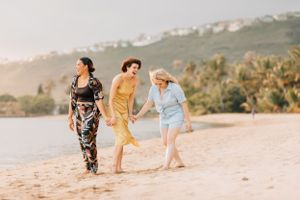 Three people in colorful dresses walk along a sunny beach with mountains in the background.