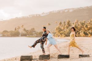 Three people jumping playfully between concrete posts on a tropical beach at sunset.