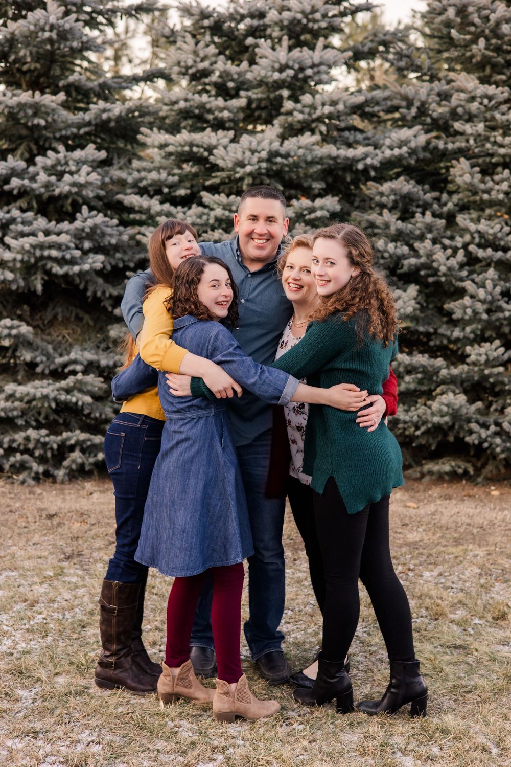 Happy family group hugging and laughing together in front of snow-dusted evergreen trees.