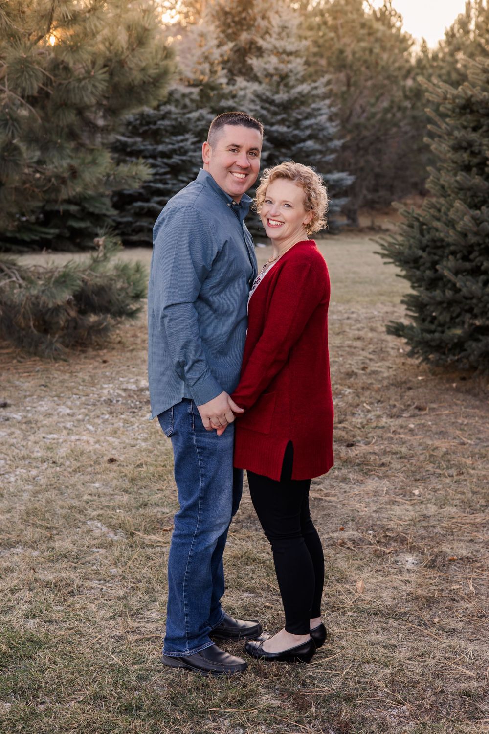 Couple in blue and red sweaters pose together among evergreen trees at sunset.
