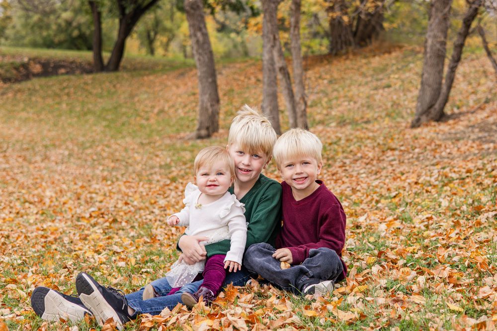 Three children sitting together on fallen autumn leaves in a park setting.