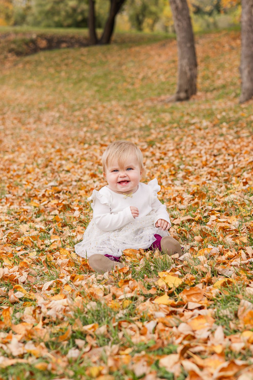 Baby in white outfit sitting among colorful autumn leaves outdoors.