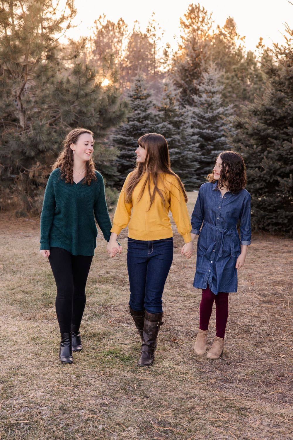 Three women in colorful sweaters walking together through pine trees at dusk.