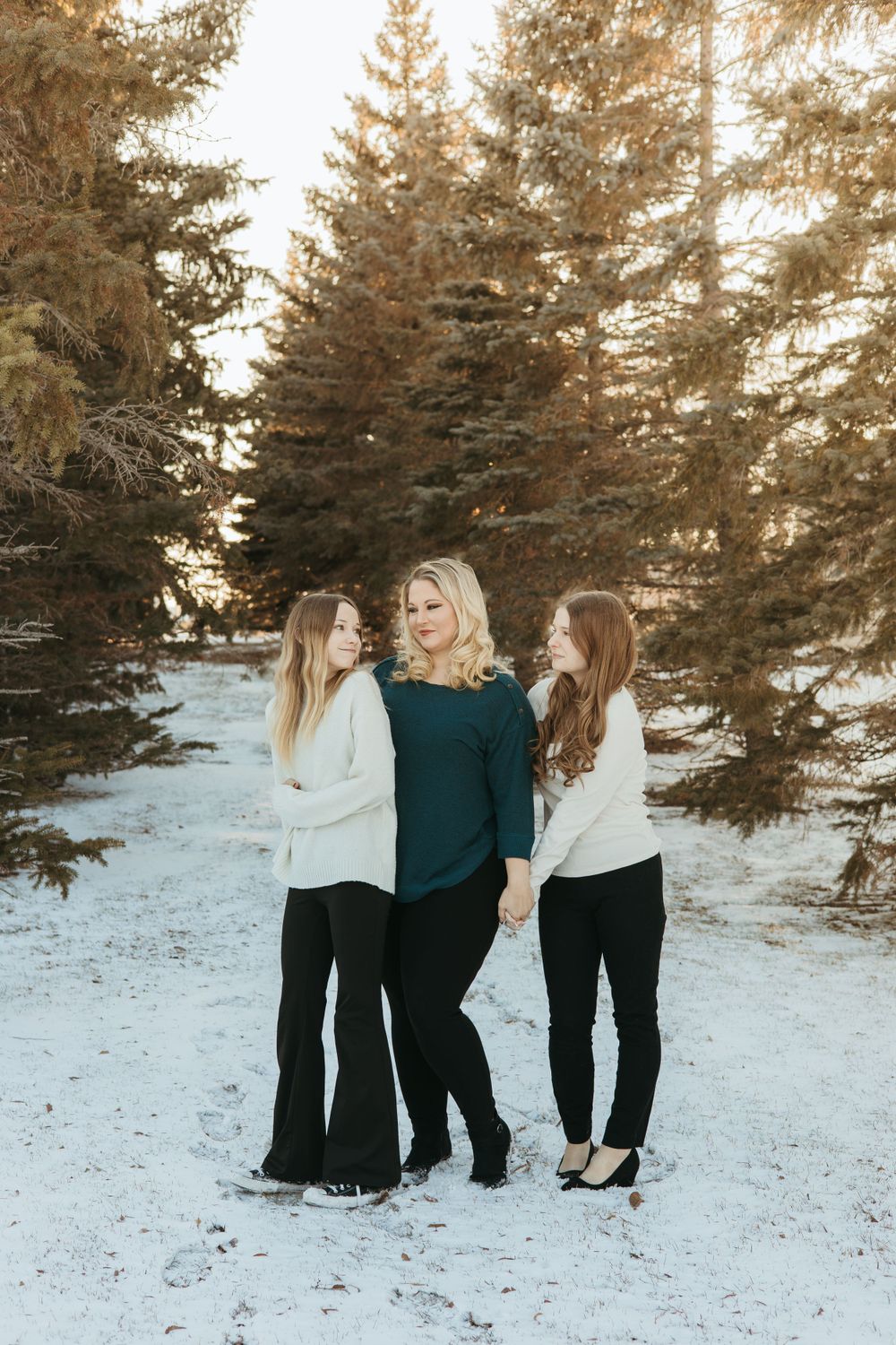 Three women standing together in winter scene with snow-covered evergreen trees.