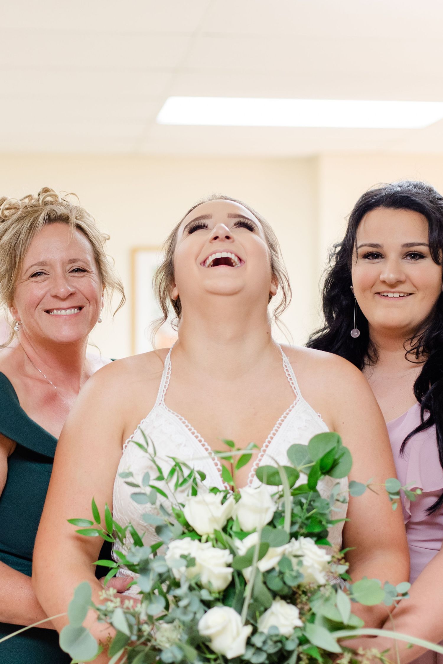 Group of bridesmaids holding white and green bouquets share joyful laughter.