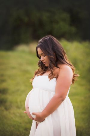 Pregnant woman in a green field with a white flowing maternity dress.  Dress has a slit and her belly is peeking through