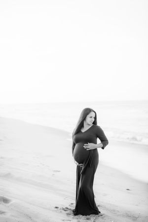 Black and white image of pregnant woman standing on a beach, looking out at the ocean.