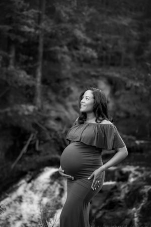 Black and white image of pregnant woman in fitted dress looking towards the sky with waterfall in the background.