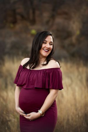 Pregnant woman in burgundy dress in a yellow field, holding below her belly and laughing.