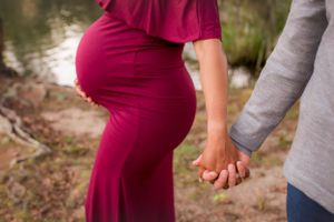 Pregnant woman's belly in a burgundy dress holding hands with her husband, Savannah River in the background.