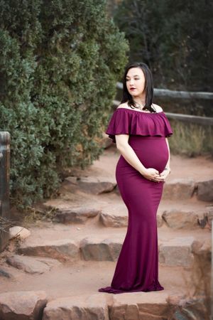 Pregnant woman in burgundy dress standing on rock steps with green shrubs.  Holding below her belly and looking off.
