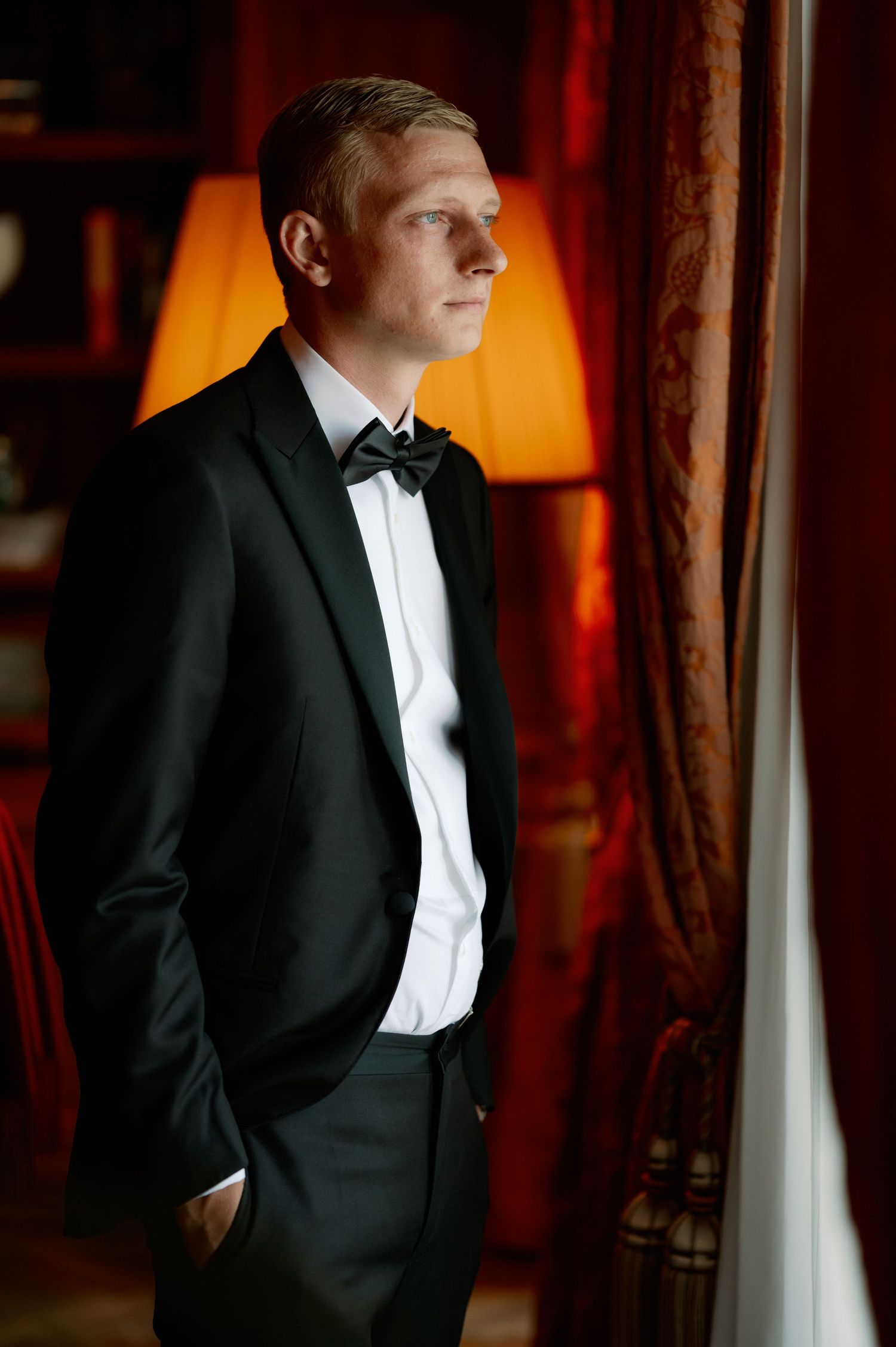 A formal portrait in warm lighting near a window with dramatic red curtains.