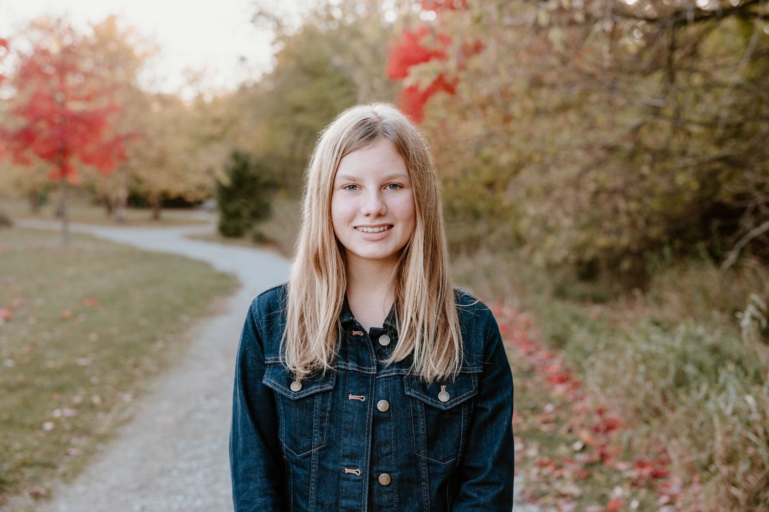 A person in a denim jacket smiles while standing on a path with red fall foliage in the background.
