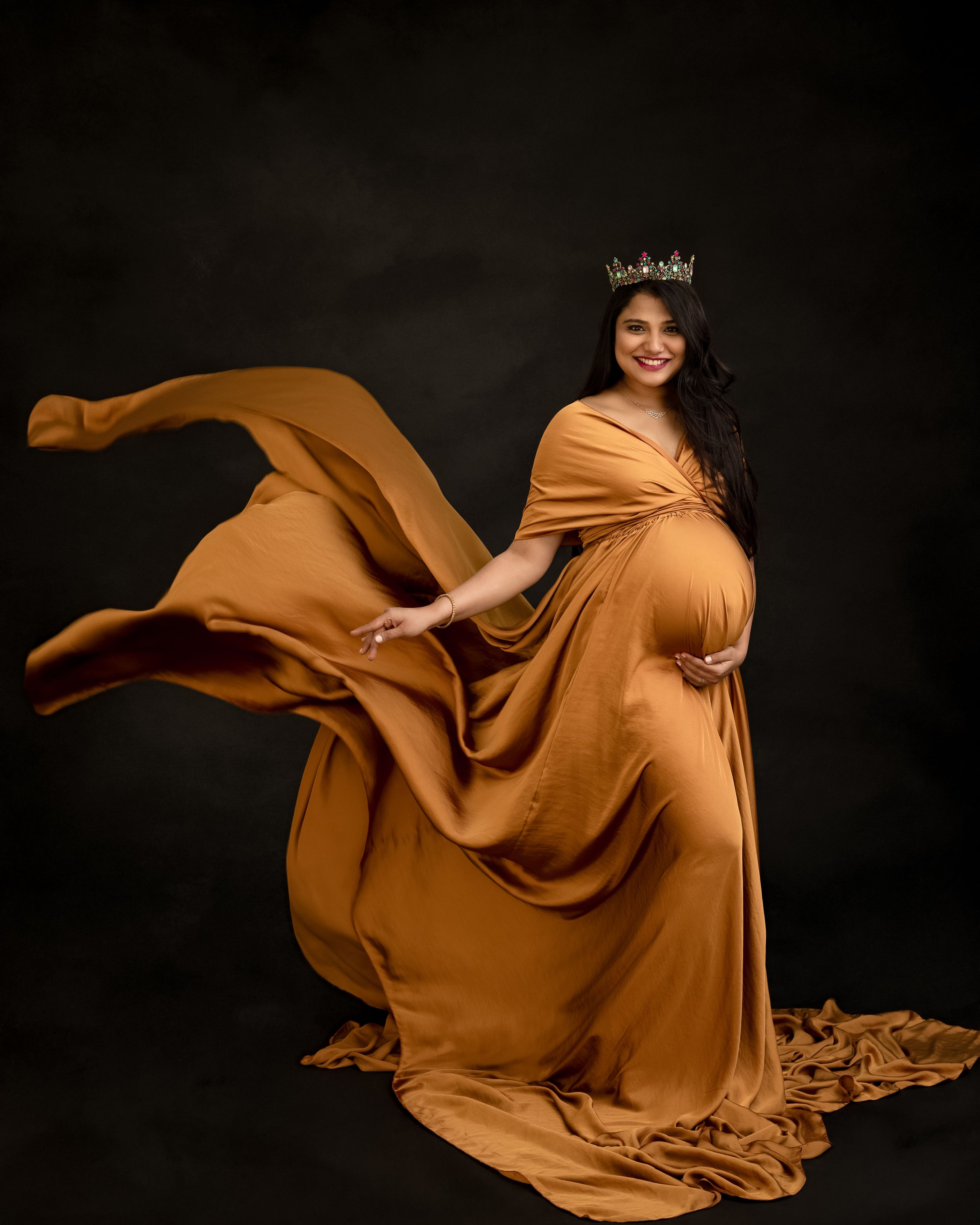 A maternity photo featuring a flowing caramel-colored dress against a dark background with dramatic fabric movement.