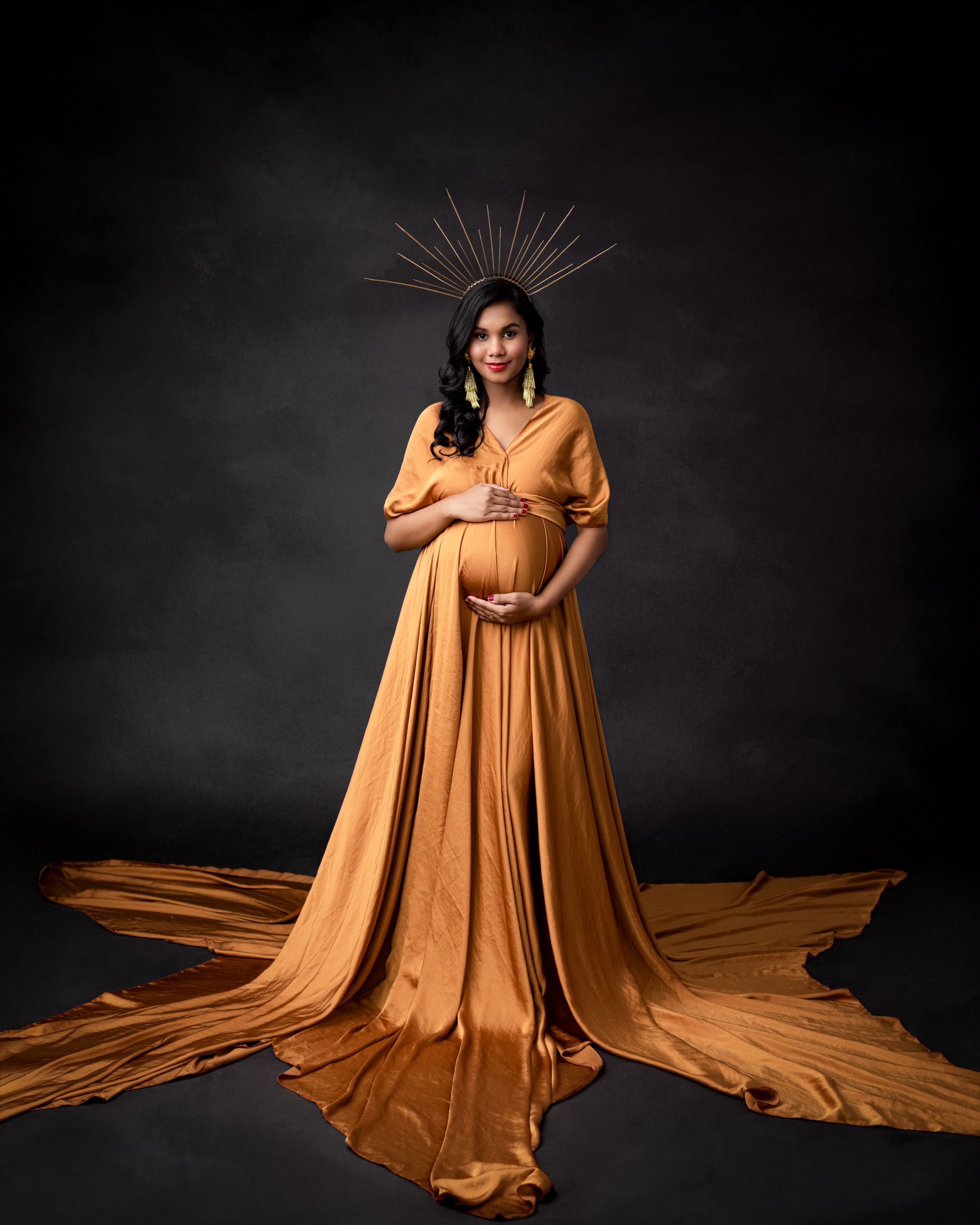 Artistic maternity portrait in flowing mustard gown with sunburst crown against dark background.
