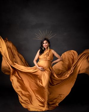 Dynamic maternity portrait with flowing orange fabric and sunburst crown against dark background.