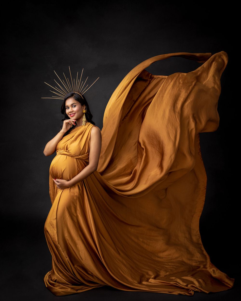 Dramatic maternity photo with billowing orange dress and metallic crown against dark backdrop.