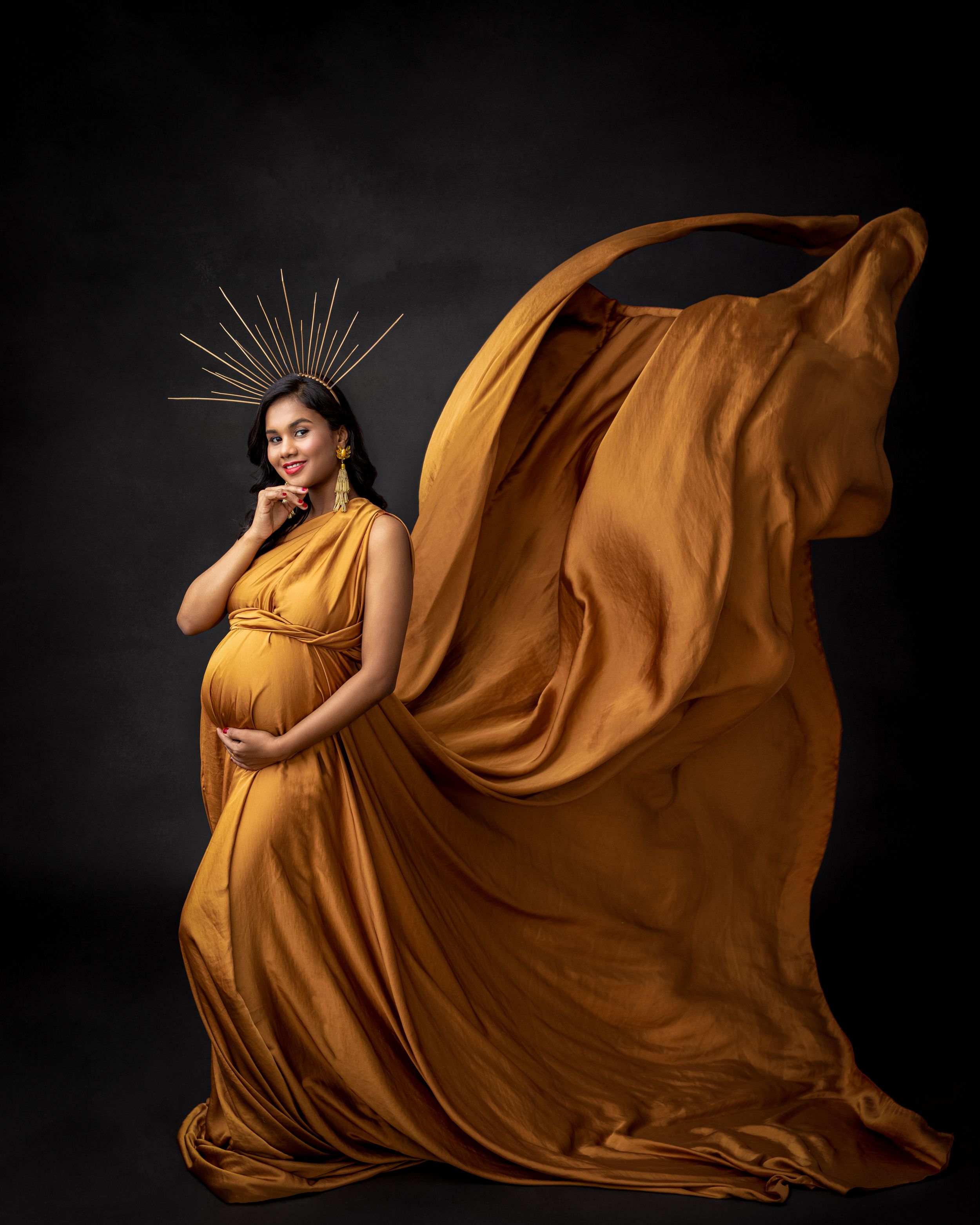 Dramatic maternity photo with billowing orange dress and metallic crown against dark backdrop.