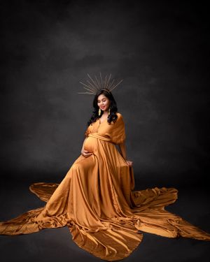 Elegant maternity portrait featuring golden dress and crown against moody dark backdrop.
