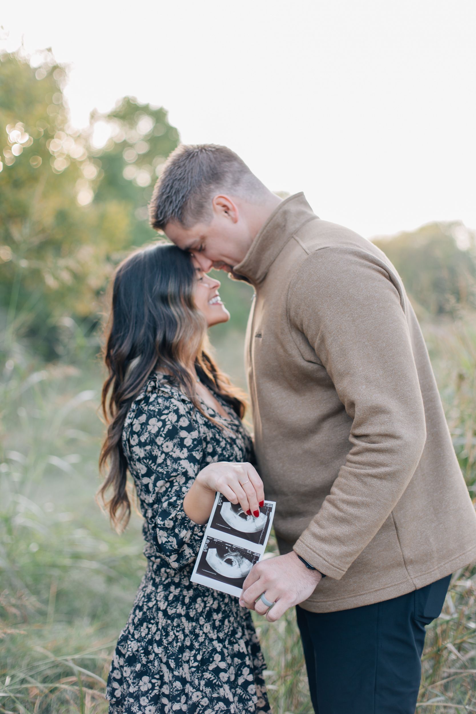 Couple embracing outdoors while holding ultrasound photo during pregnancy announcement.