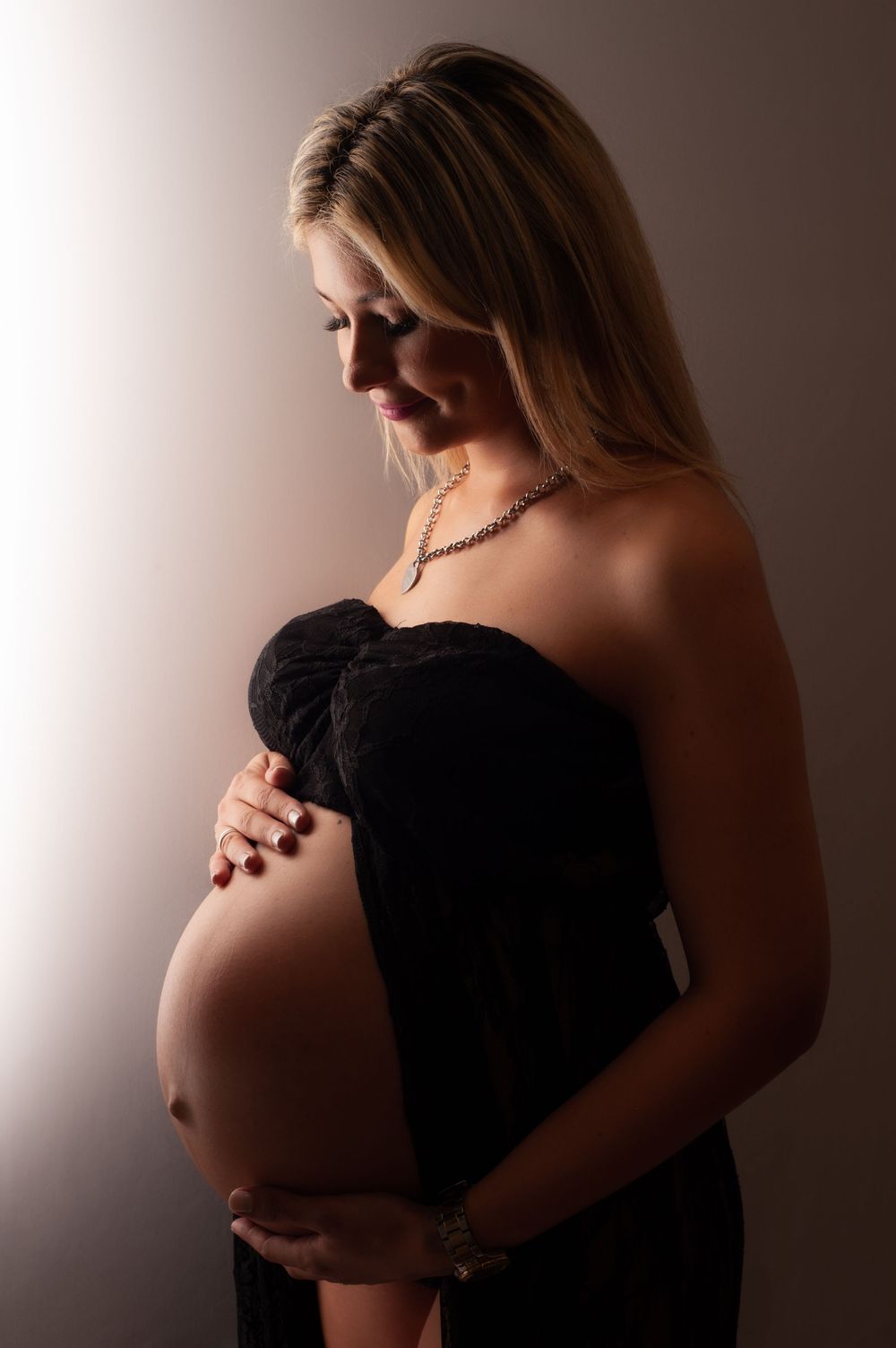Elegant pregnant woman in black strapless dress holding baby bump in dramatic studio lighting.